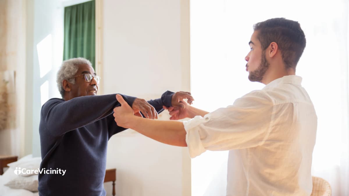 A support worker helping an older man with a gentle arm exercise in a bright home bedroom