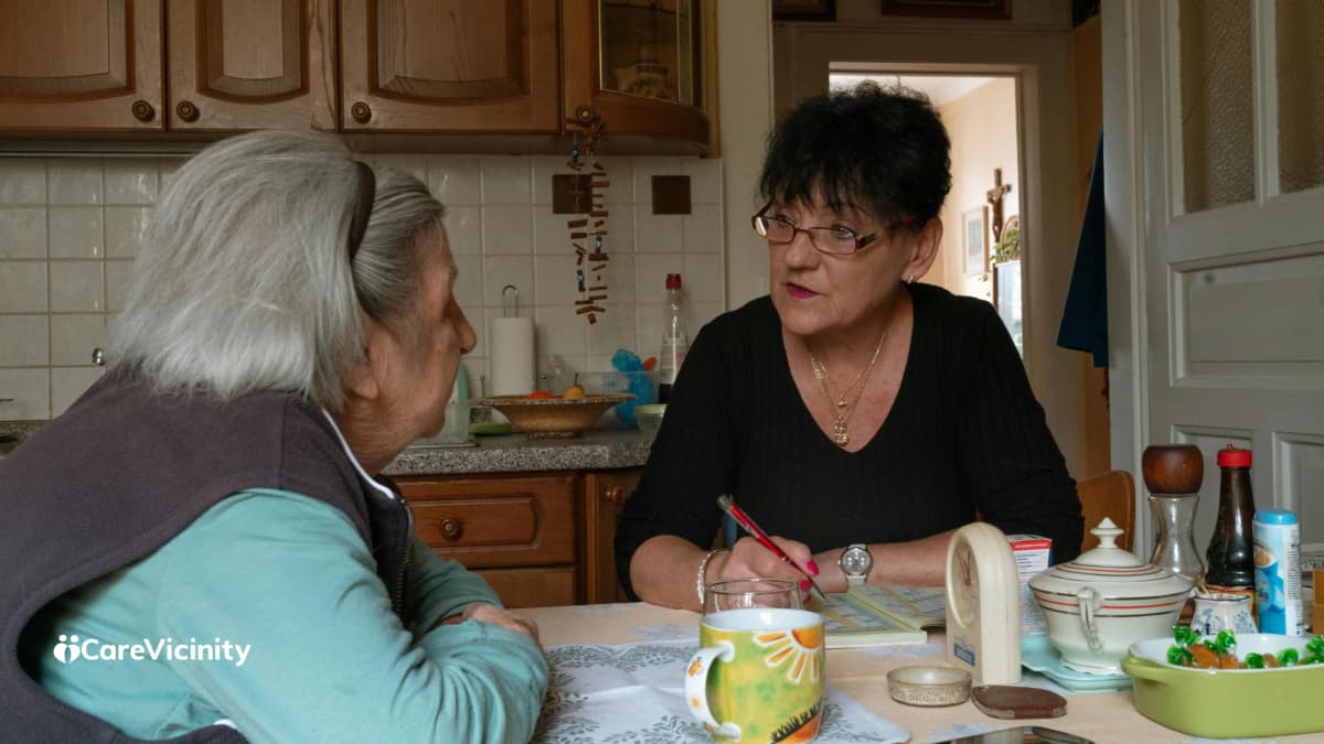 Two women sitting at a kitchen table talking and taking notes together.