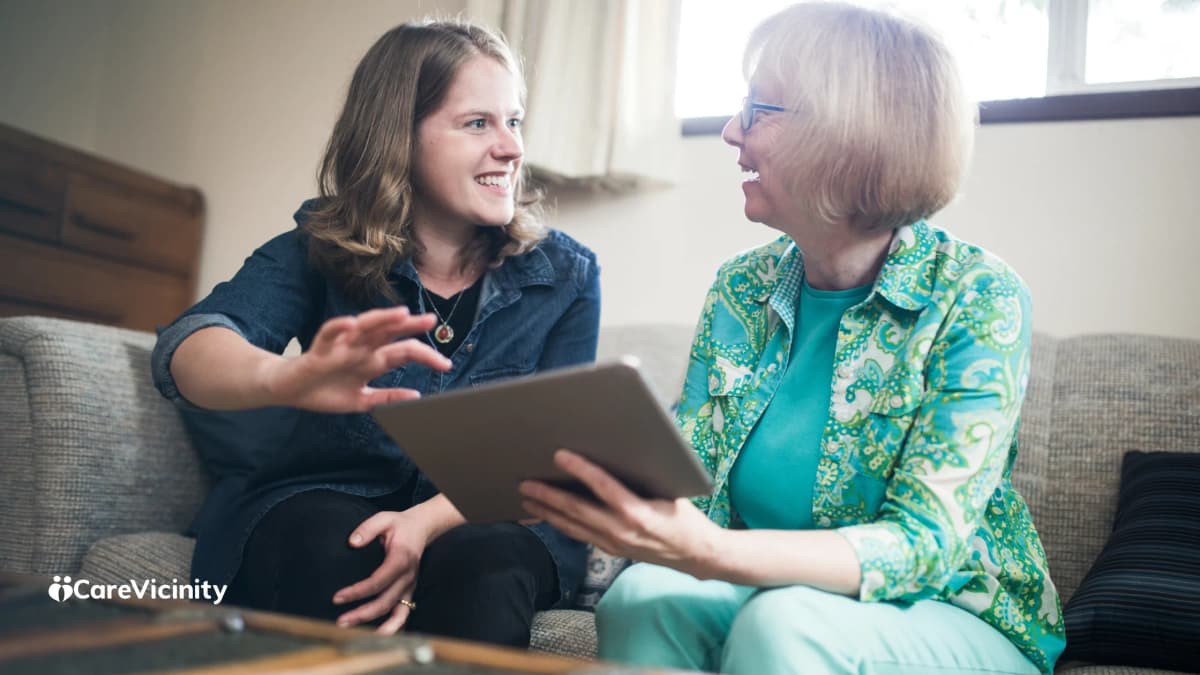 Older woman and a younger woman laughing together on a sofa at home while looking at a tablet