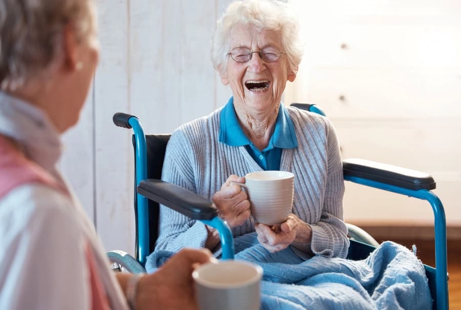 Older woman in a wheelchair laughing while holding a mug indoors