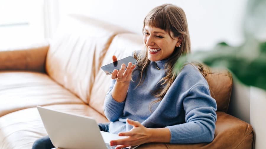 Woman smiling while speaking into smartphone with laptop on couch