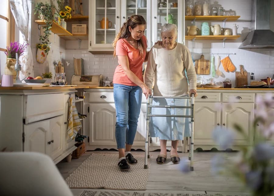 Younger woman assisting an older woman using a walking frame in the kitchen