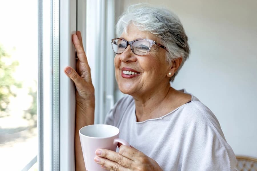 Older woman smiling while holding a mug by a window