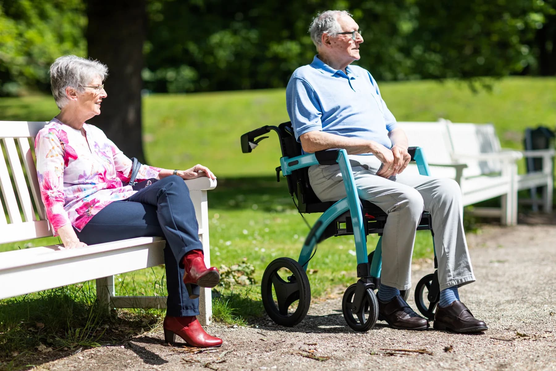 Older man resting on his rollator in a sunny park beside an older woman on a bench