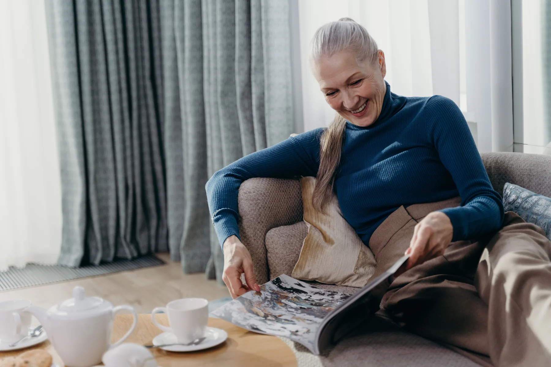 Older woman smiling as she reads a magazine over a pot of tea at home