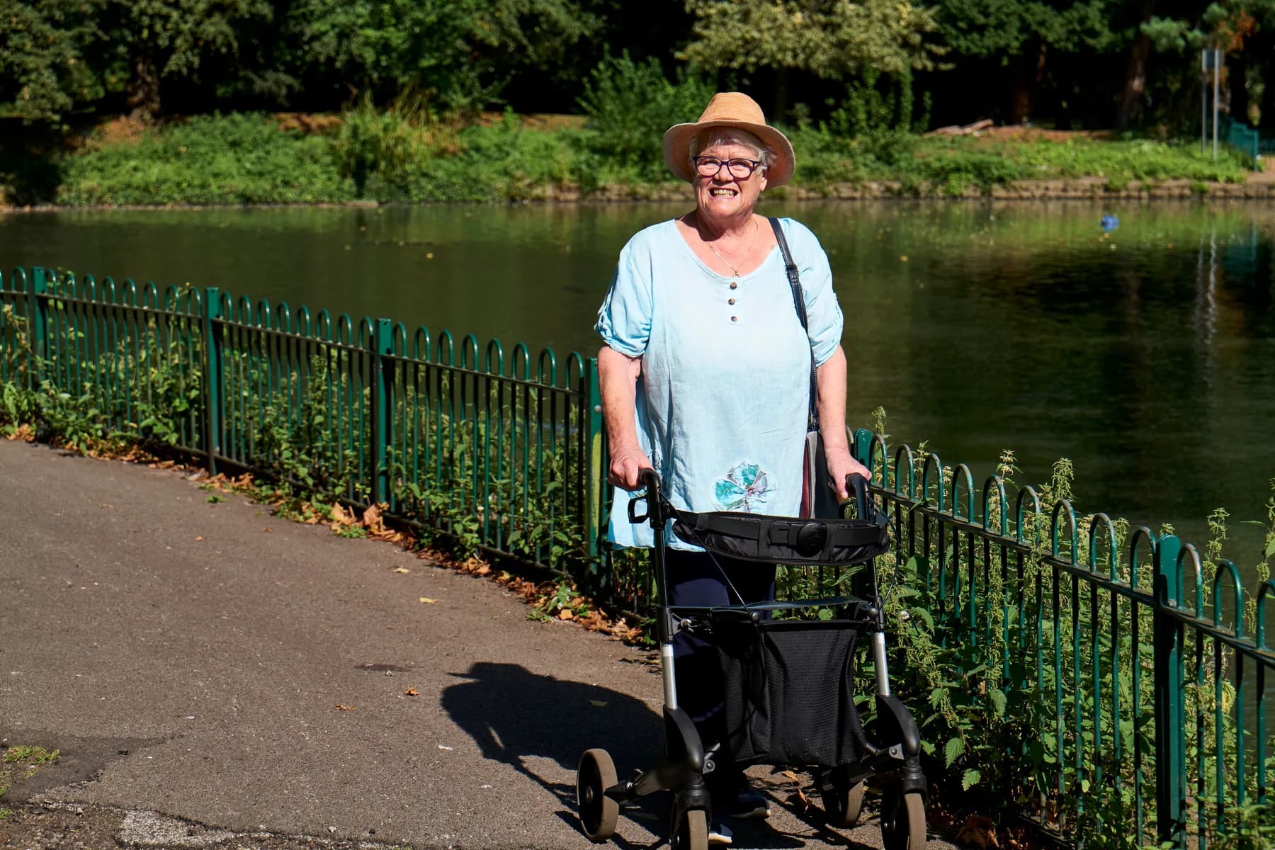 Older woman smiling as she walks with her walking frame along a sunny park path