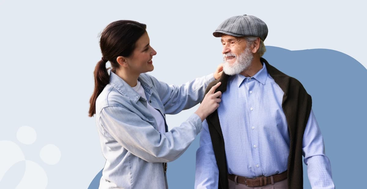 A woman in a denim jacket smiles and adjusts the jacket of an older man in a flat cap against a light blue background.
