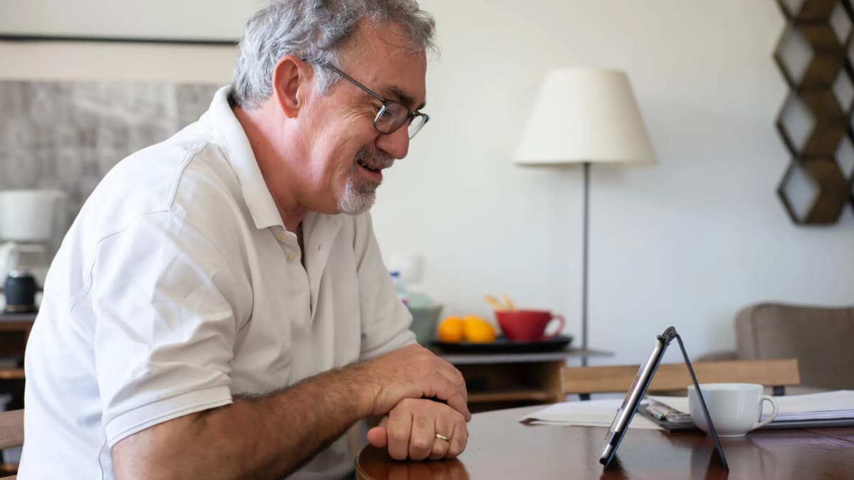 An older man smiling as he talks on a video call on a tablet at his dining table