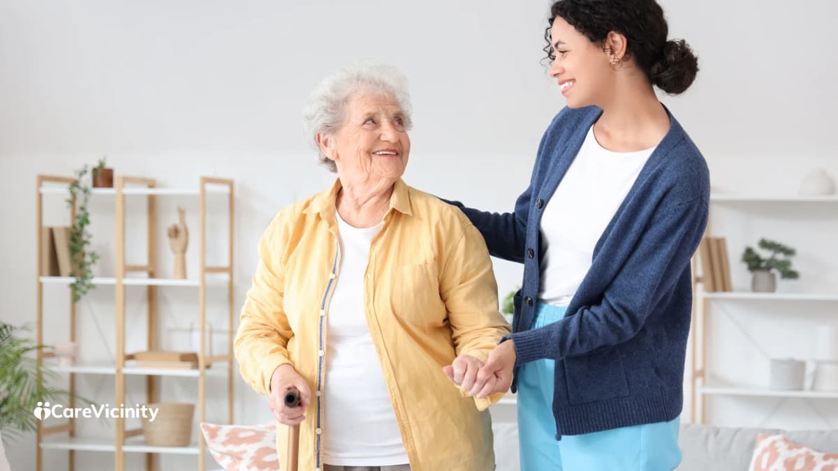 Older woman with a walking cane smiling at a support worker beside her in a bright living room