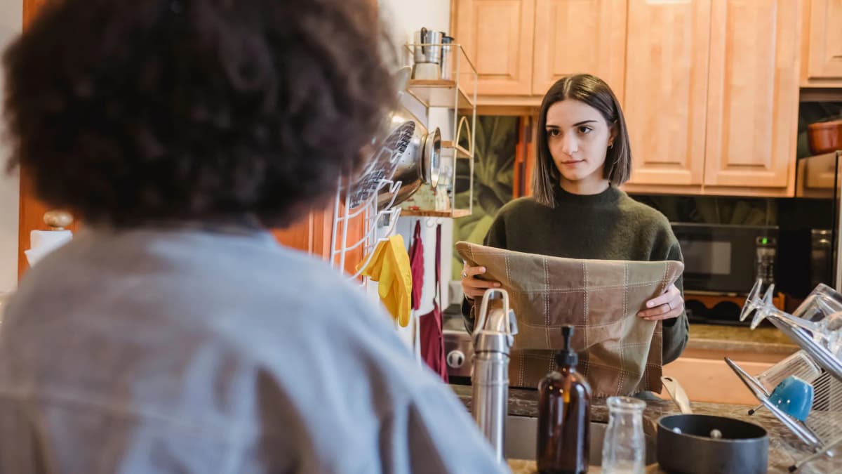 Two women having a serious talk in a home kitchen, one seen from behind and the other holding a tea towel
