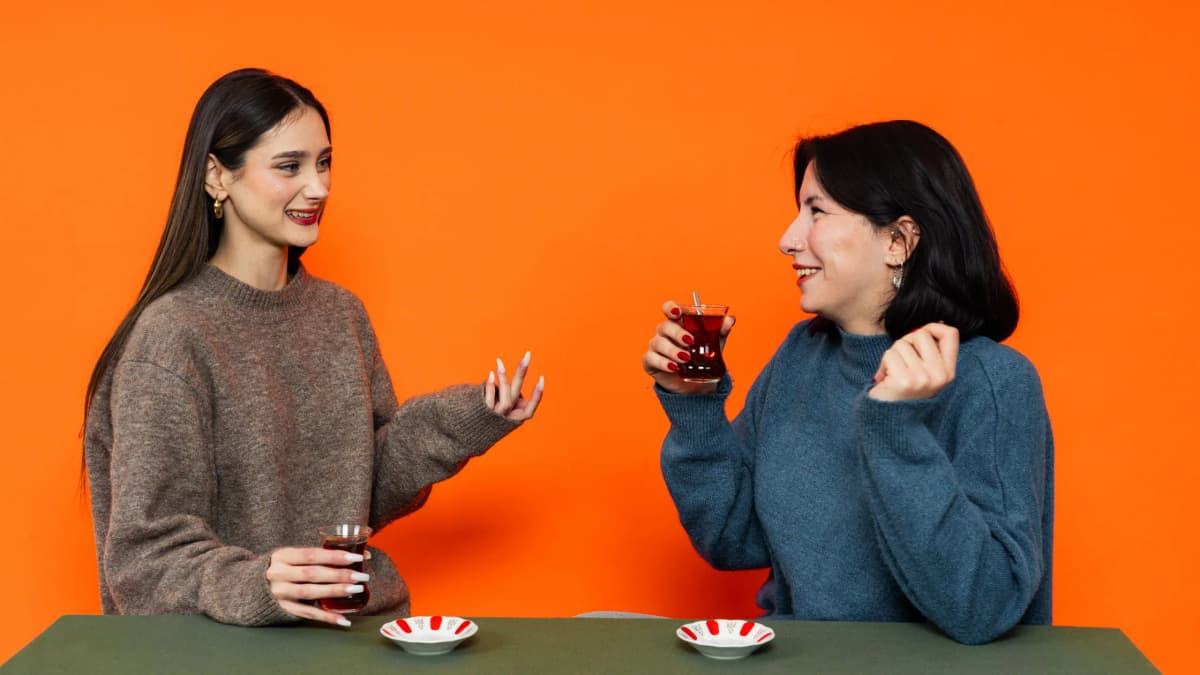 Two women smile and talk over glasses of tea at a table against an orange wall