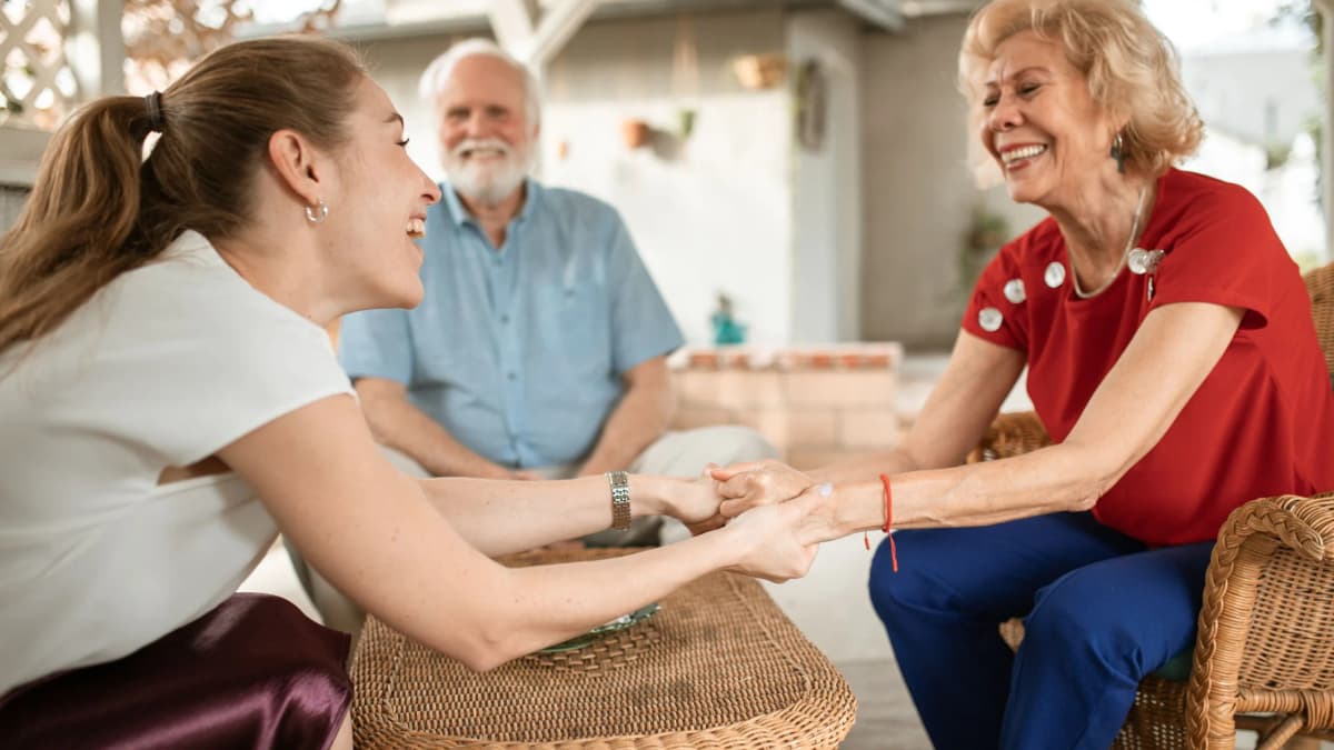 A younger woman holds hands and laughs with an older woman on a home veranda while an older man smiles behind them