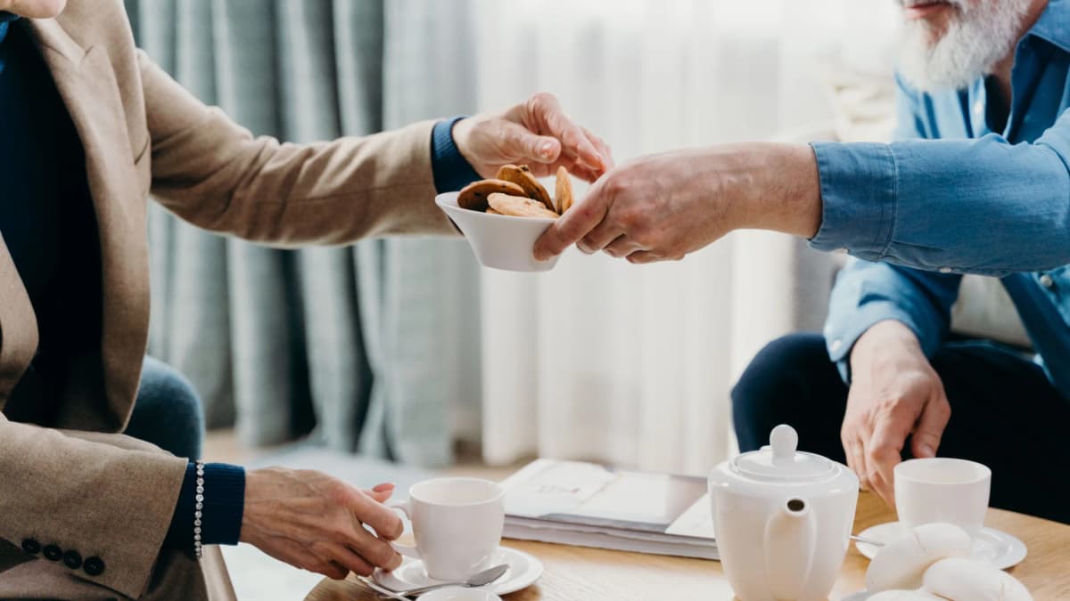 Two older adults sharing biscuits and a pot of tea at a coffee table at home