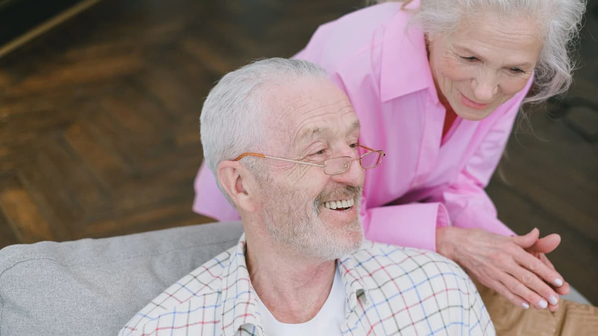 An older man smiling on a sofa at home while an older woman leans over his shoulder