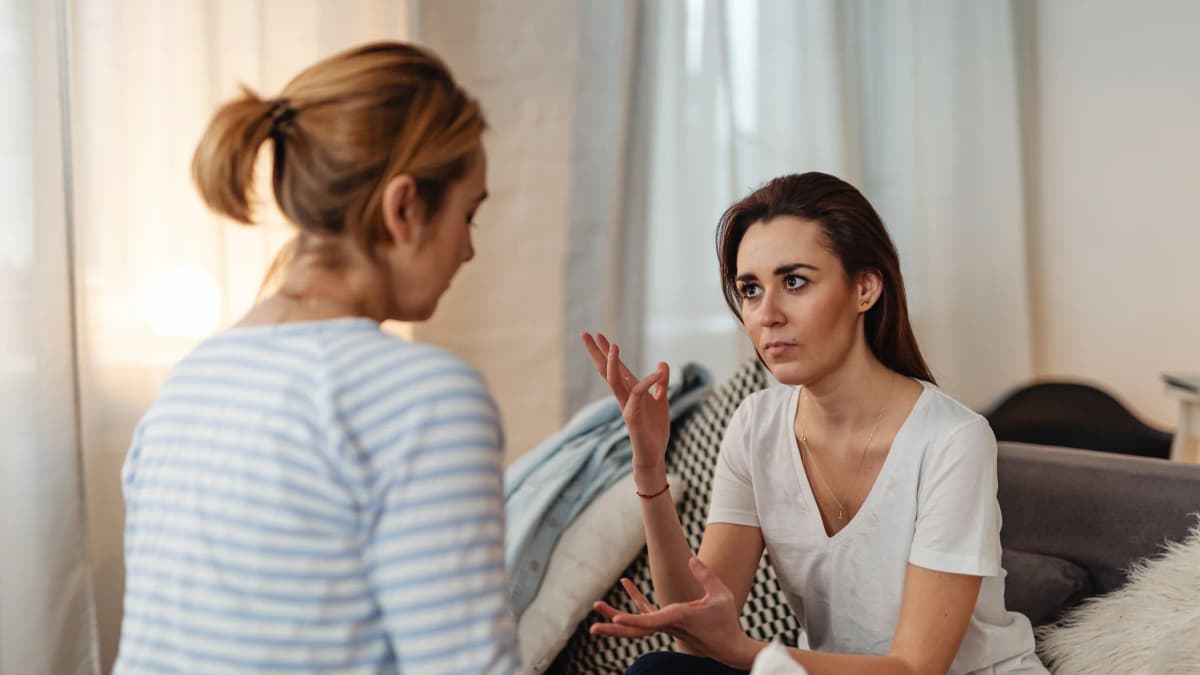 Two women sitting on a sofa at home, one talking calmly while the other listens