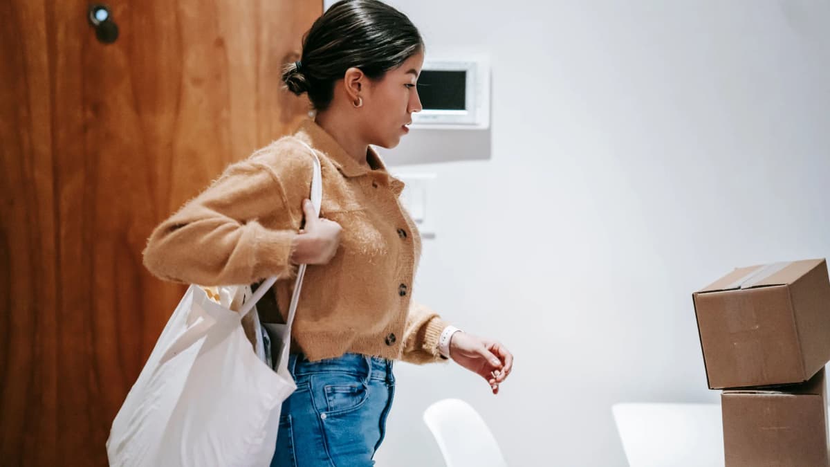 A woman with her hair tied back carries a white tote bag over her shoulder as she walks into a home