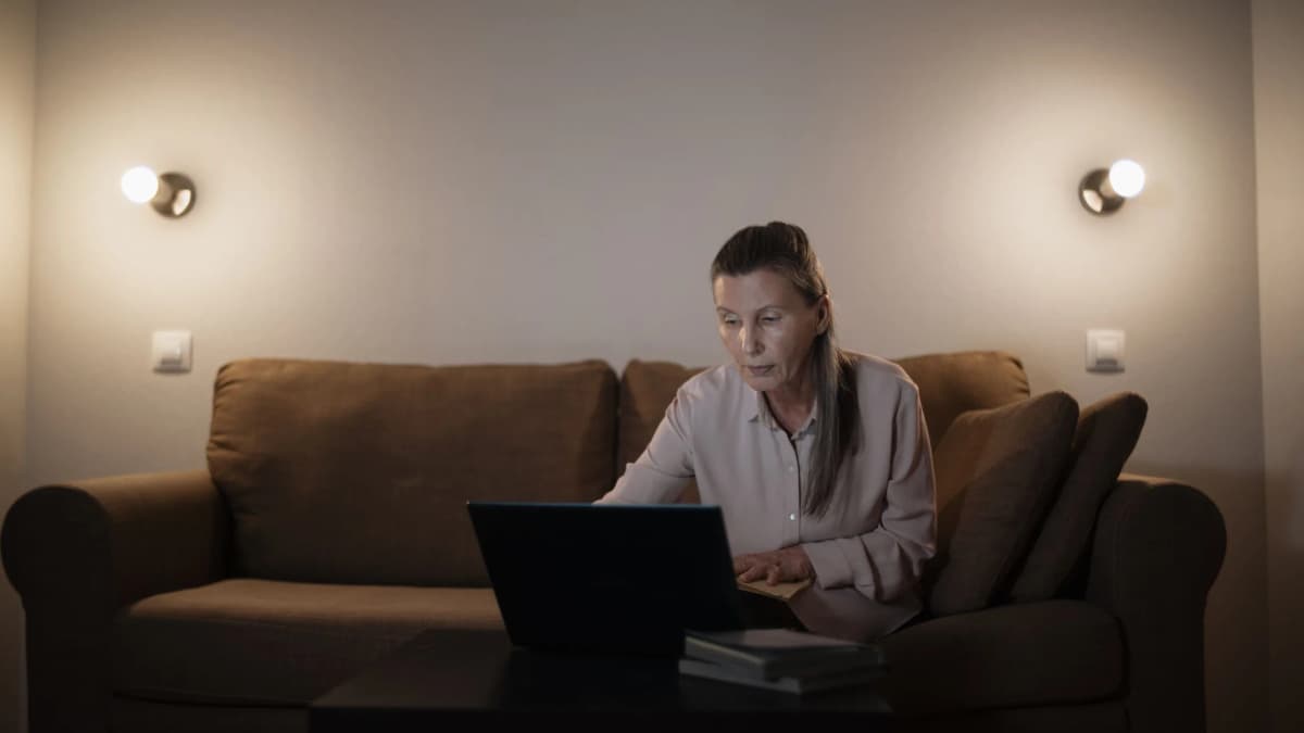 Older woman sitting on a sofa at home in the evening, using a laptop under warm wall lamps