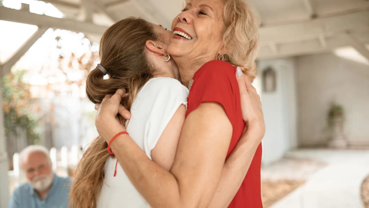 A younger woman hugging a smiling older woman on a sunny porch, with an older man watching in the background