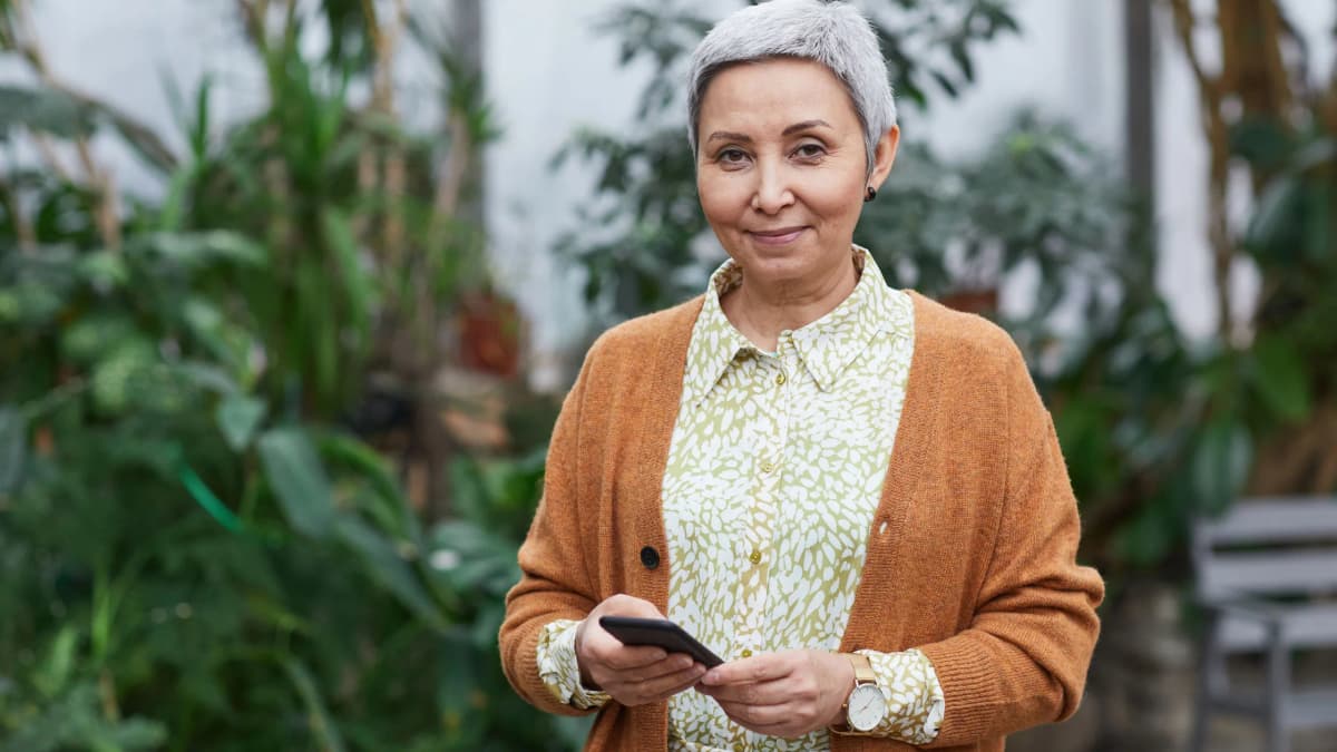 An older woman with short grey hair holding a mobile phone among indoor plants