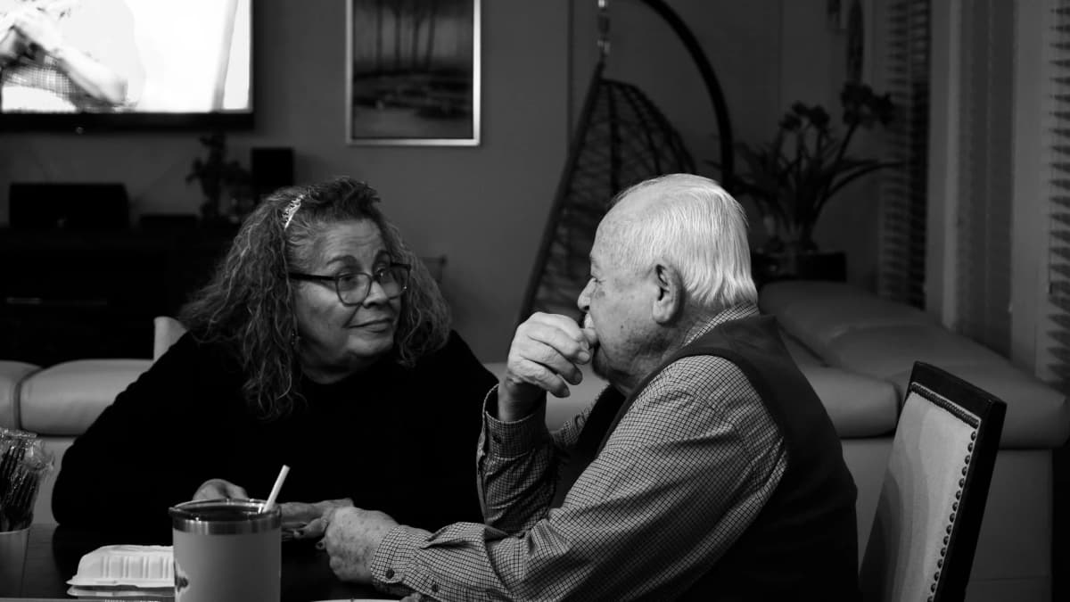 An older man and a woman talk across a dining table at home, in black and white