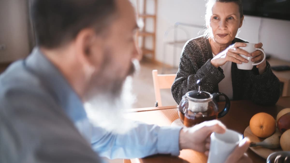 An older woman holds a mug at a kitchen table with a teapot and fruit, talking with an older man with a grey beard