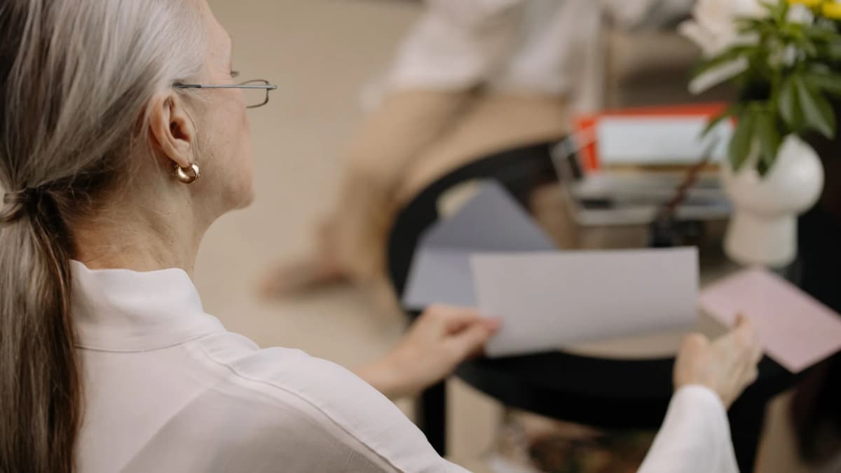 An older woman with grey hair and glasses holding a sheet of paper in her living room