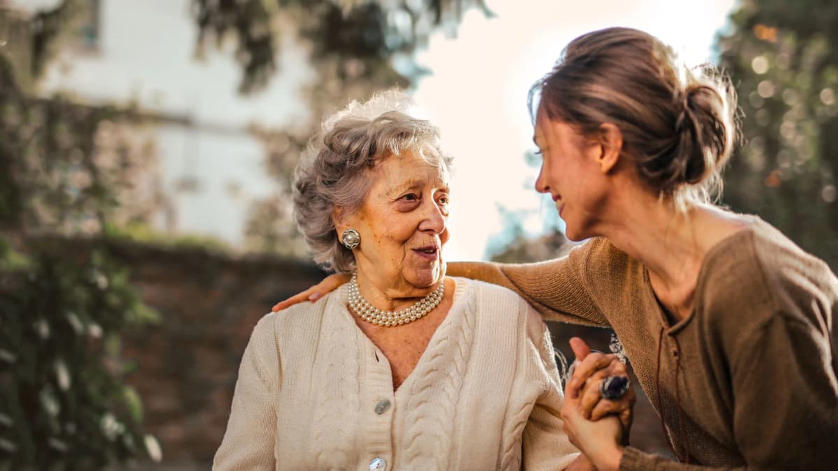 An older woman and a younger adult woman smiling at each other outdoors, holding hands