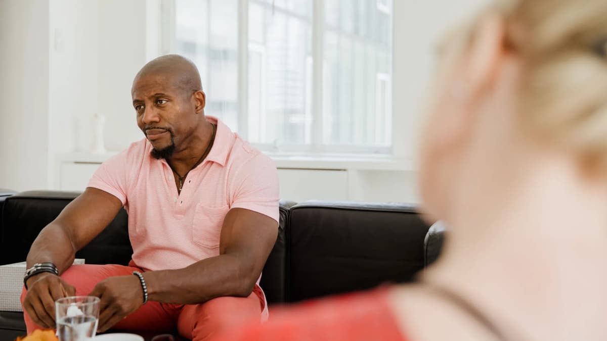 A man in a pink polo shirt sits on a sofa listening to a woman seen out of focus in the foreground