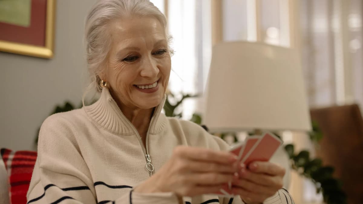 An older woman with grey hair smiles as she holds a hand of playing cards in a living room
