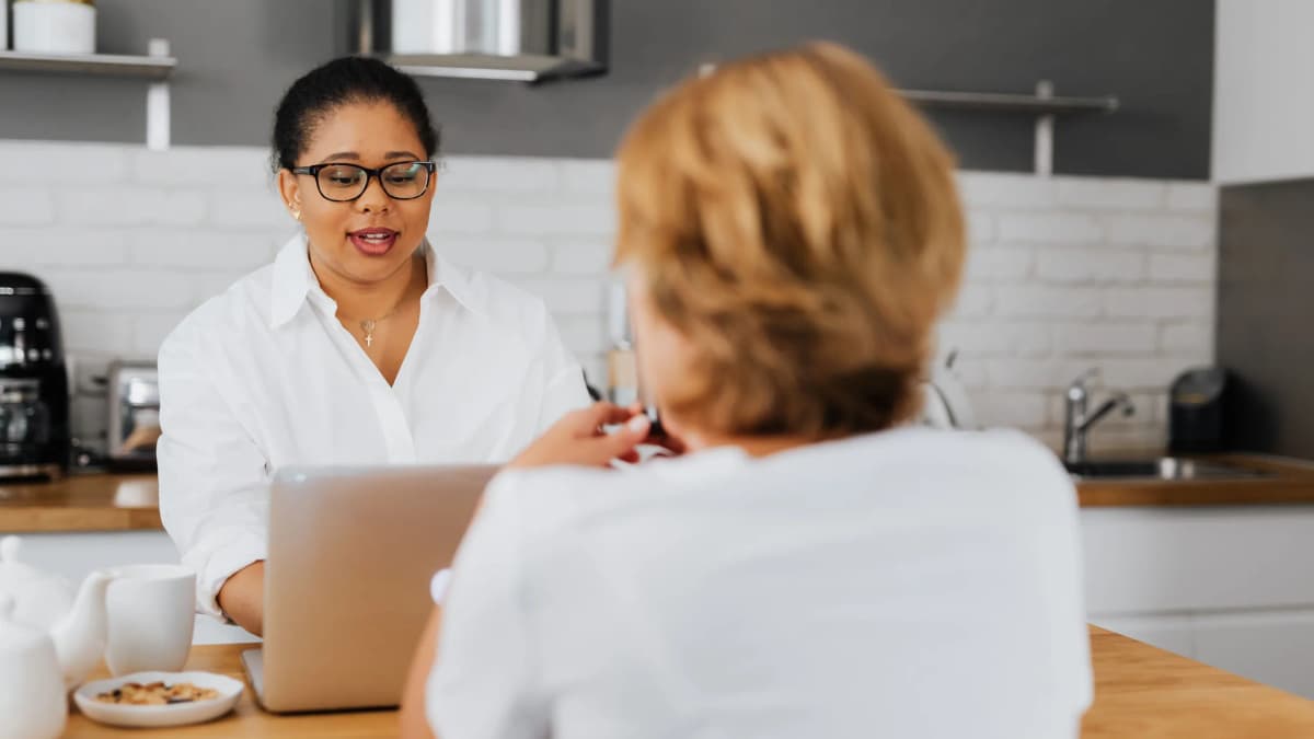 A woman in glasses talks across a kitchen table with an older woman seen from behind