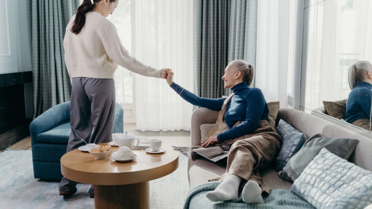 A younger woman holding hands with a smiling older woman on a sofa in a bright living room