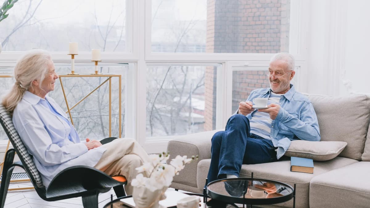 An older woman and an older man smiling and talking over a cup of tea in a bright living room