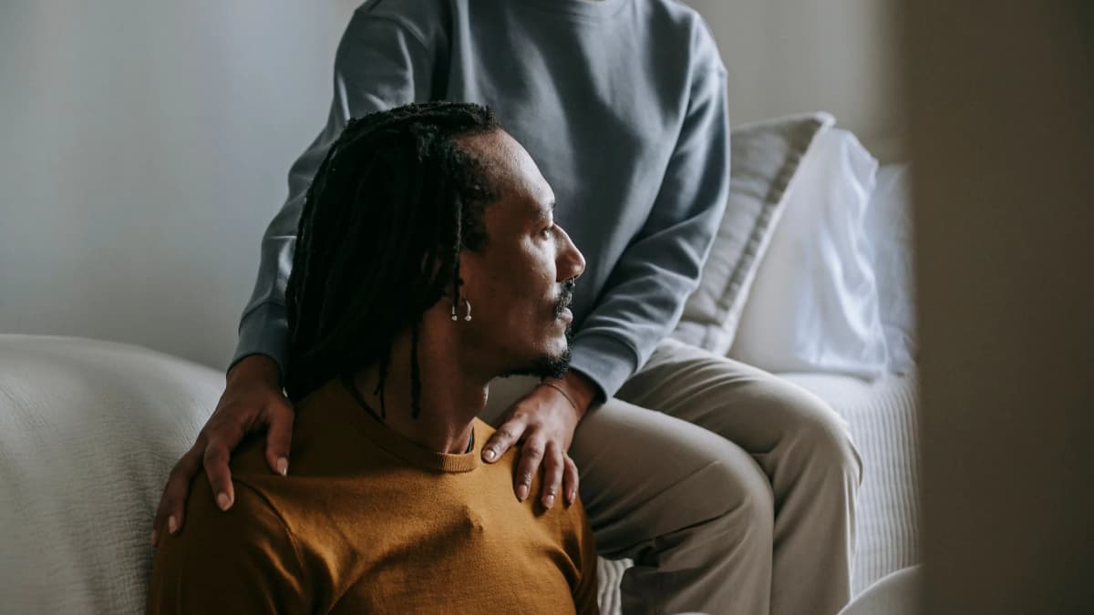A person rests their hands on the shoulders of a man with long locs who sits looking away, deep in thought