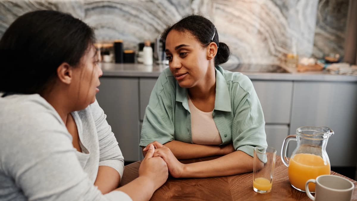 Two women talking at a kitchen table, one gently holding the other's hands