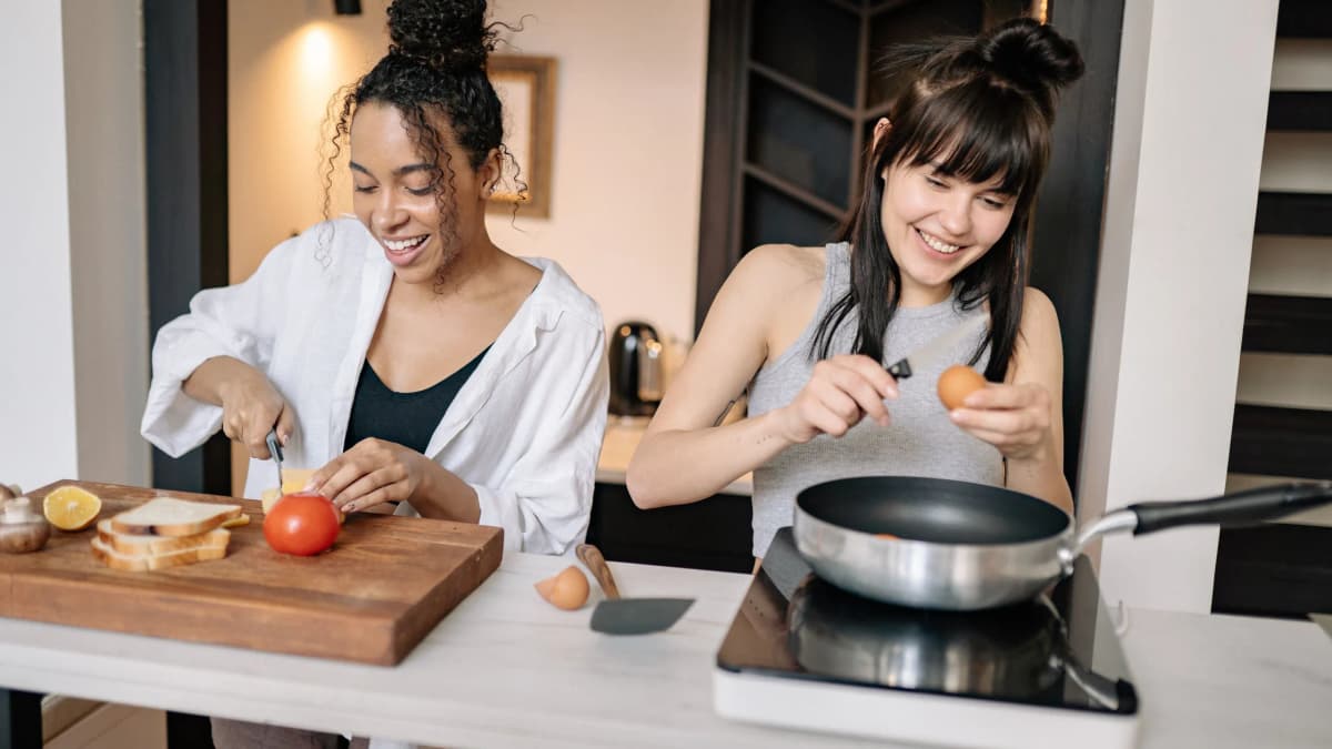 Two women preparing a meal together in a home kitchen, one slicing a tomato and one cracking an egg into a pan