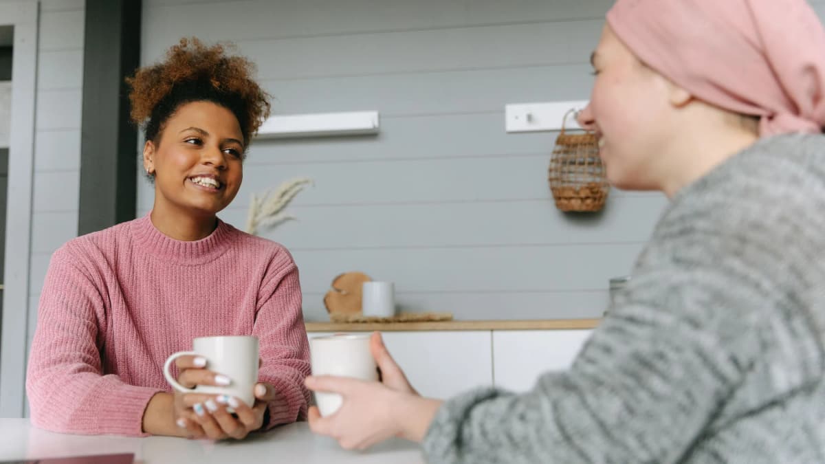 Two women smiling and talking over mugs of tea at a kitchen table at home
