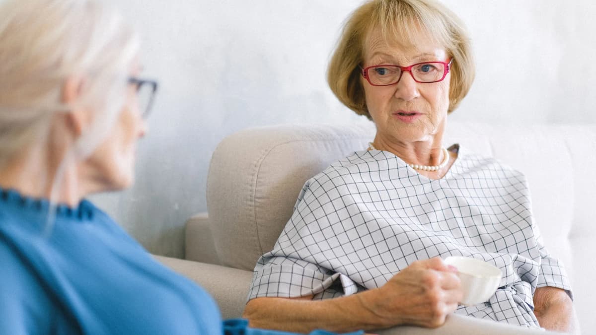 Two older women talk in a bright living room, one holding a white cup and listening