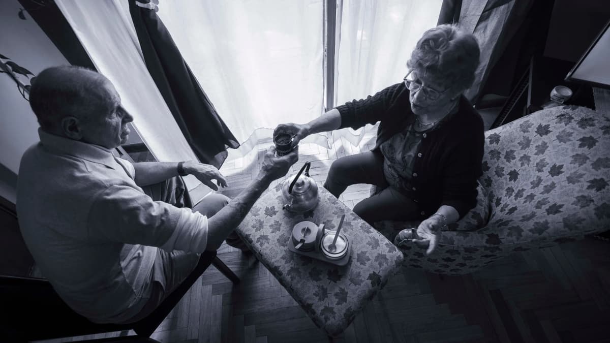 An older man and woman sitting by a window at home, sharing a hot drink and talking