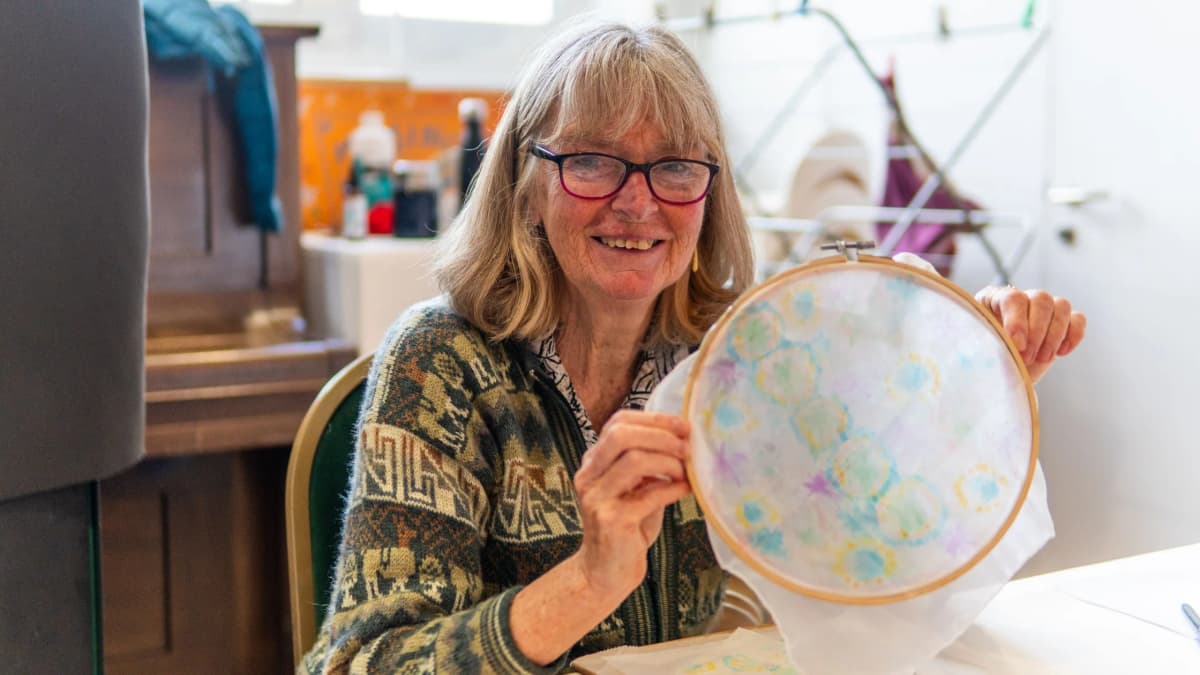 An older woman with glasses smiling as she holds up a piece of her own embroidery in a wooden hoop