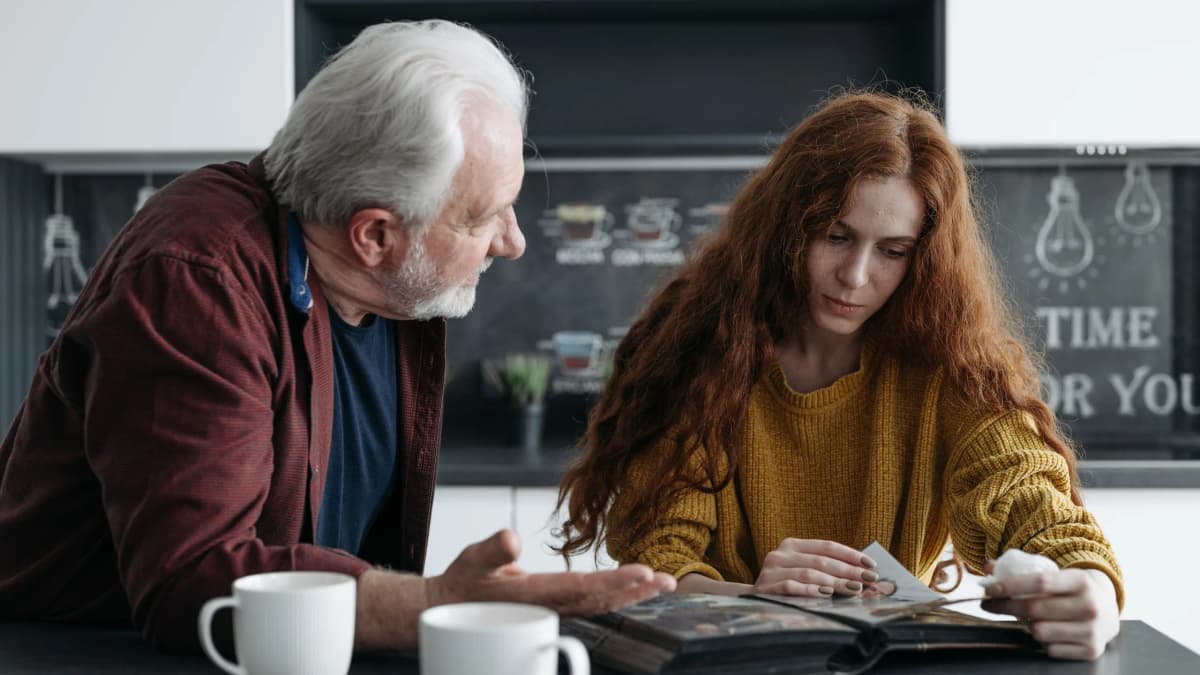 An older man speaks seriously to a younger woman with a photo album at a kitchen bench