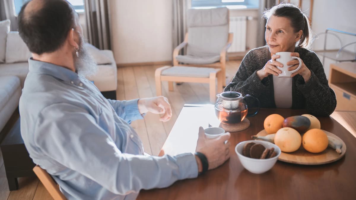 An older man and woman talking over tea at a kitchen table at home