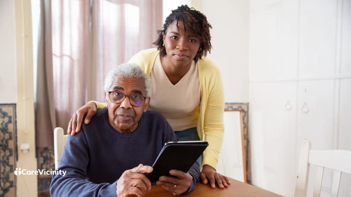 An older man at a table with a tablet, a younger support worker standing behind him with a hand on his shoulder