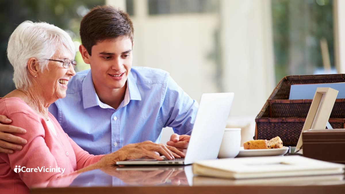 A young man and an older woman sitting together at a table, smiling, looking at a laptop