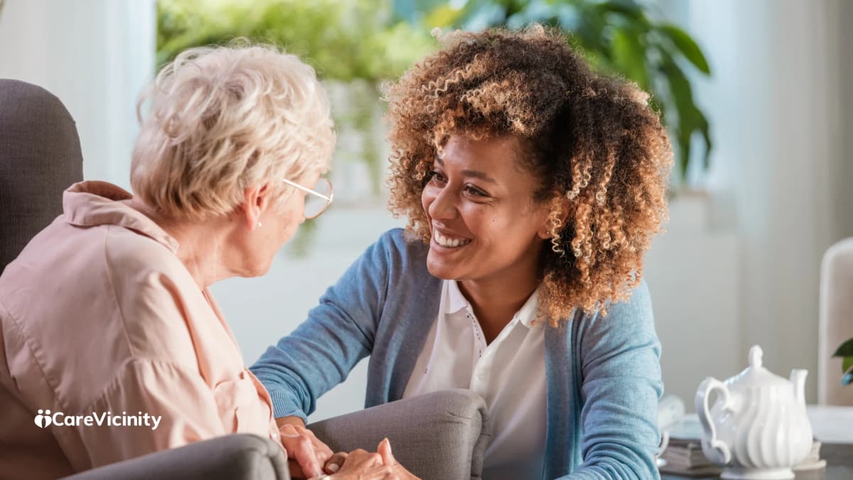 Two women sitting together indoors, smiling warmly in conversation.