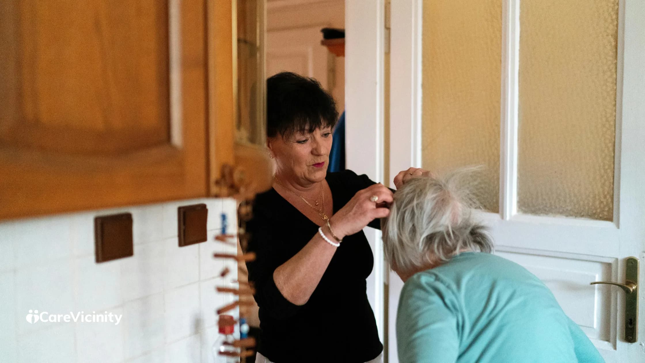 Support worker helping an older woman with her hair in a home kitchen