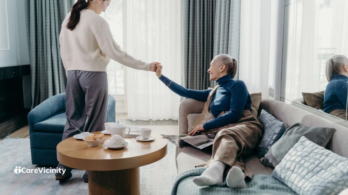 A support worker and an older client holding hands warmly in a bright living room, tea on the table