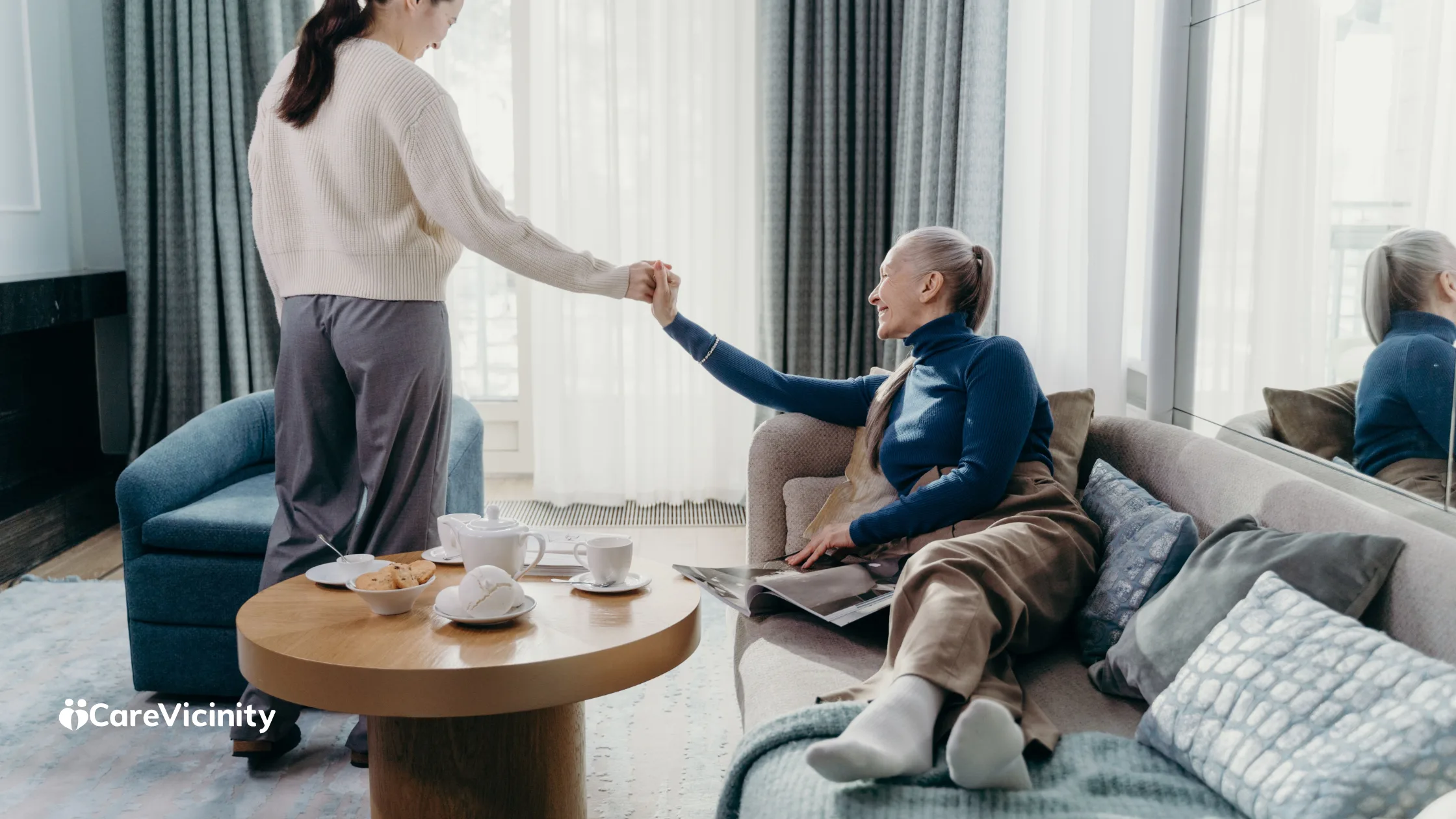 A support worker and an older client holding hands warmly in a bright living room, tea on the table