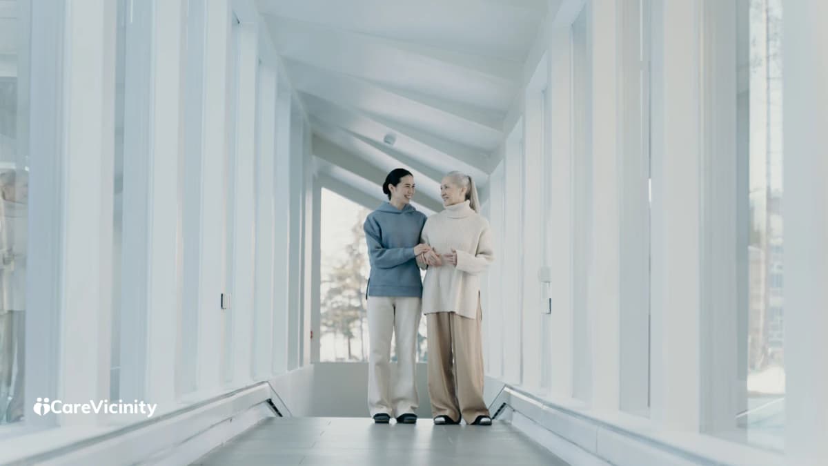 A support worker walking arm-in-arm with an older woman along a bright glass-walled hallway