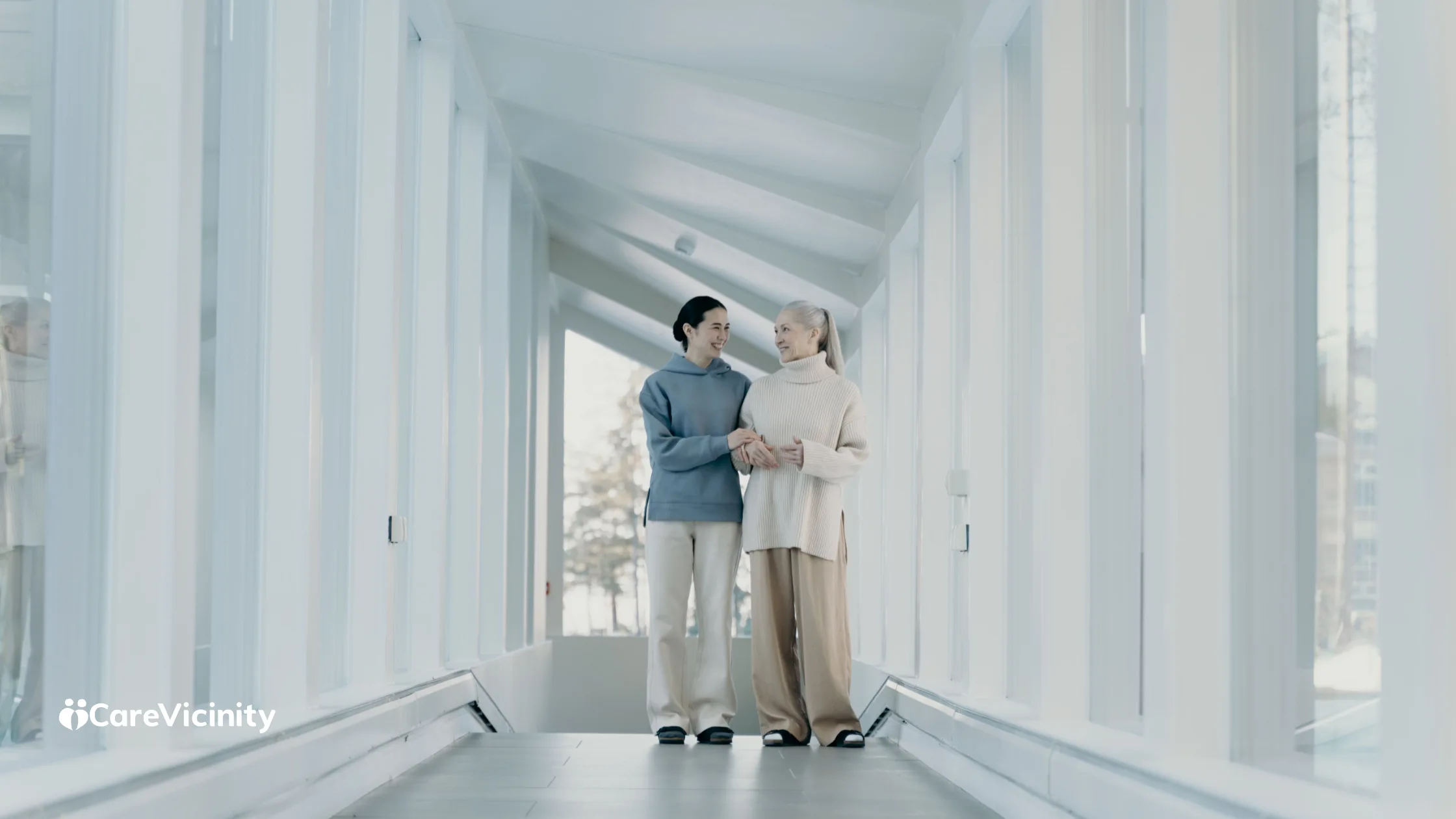 A support worker walking arm-in-arm with an older woman along a bright glass-walled hallway