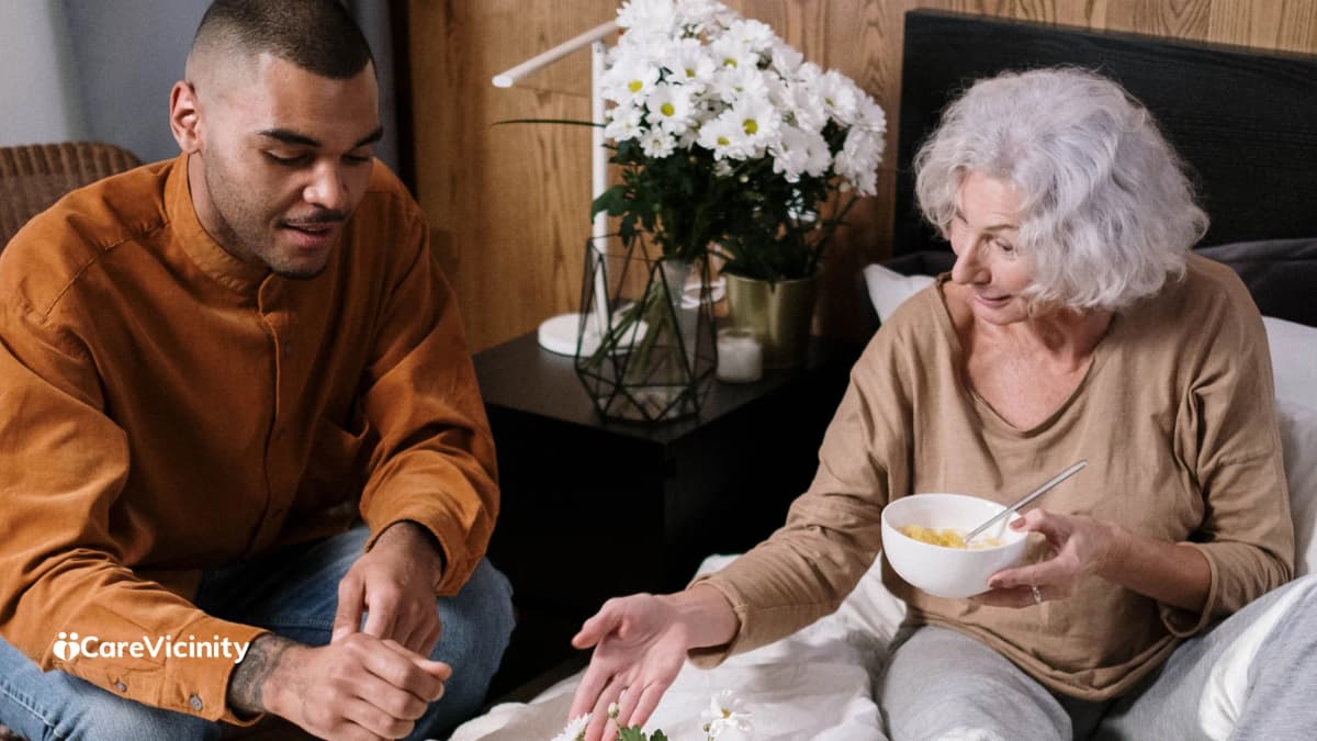 A support worker helping an older woman with a meal in a warm bedroom setting